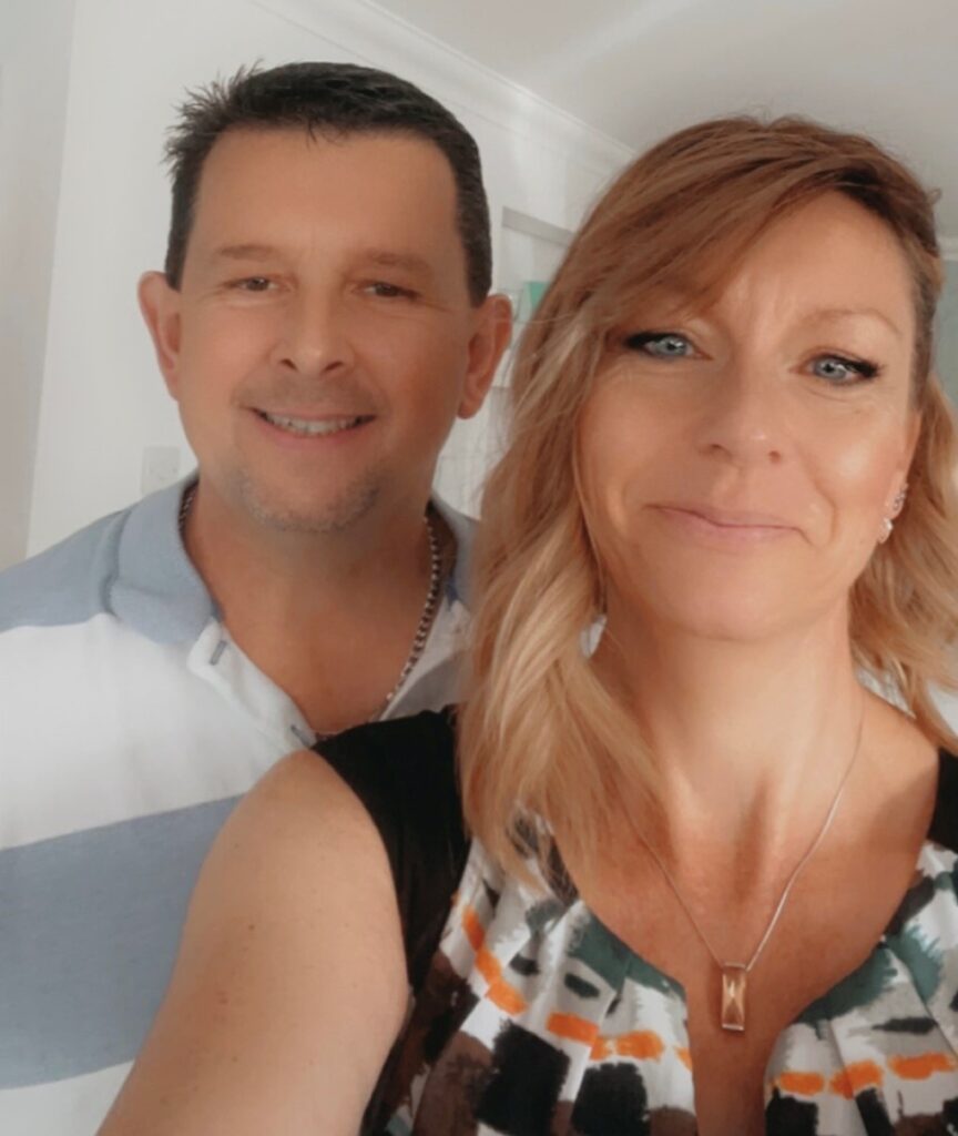 The photo shows a selfie of a woman and man from just below the shoulders. The woman in forefront is wearing a dark top with colourful lapel and has brown hair, the man is wearing a light blue top and has dark hair.