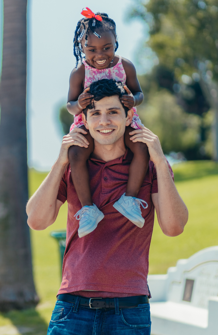 The image shows a man smiling looking to camera in a park environment with a young girl sat on his shoulders