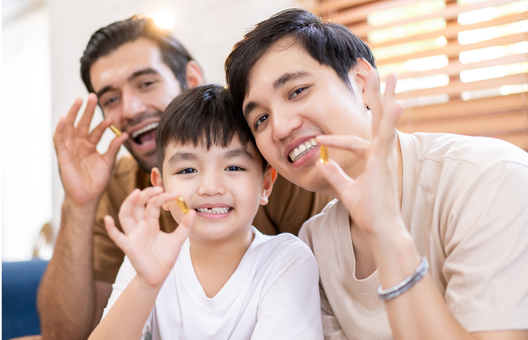You see two men and a young boy sat on a sofa smiling looking at camera holding a cod liver oil capsule.