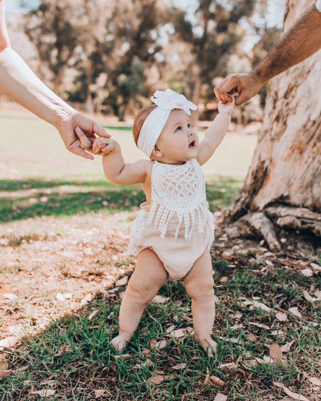 The image shows a young baby in a white romper with a white headband on holding the hands of two adults who you can't see whilst walking in a park environment.