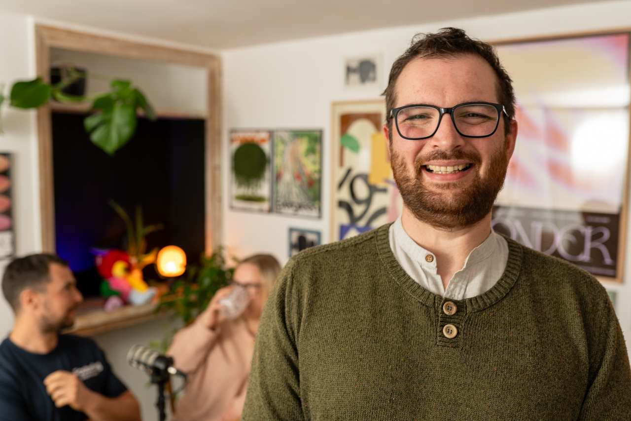 This image shows Joe, a foster carer for Wiltshire Council smiling to camera wearing a dark green jumper, with dark hair and glasses. In the background you can see a recording studio and a man with dark hair and a blue shirt looking at a woman with blonde hair whose drinking from a mug.