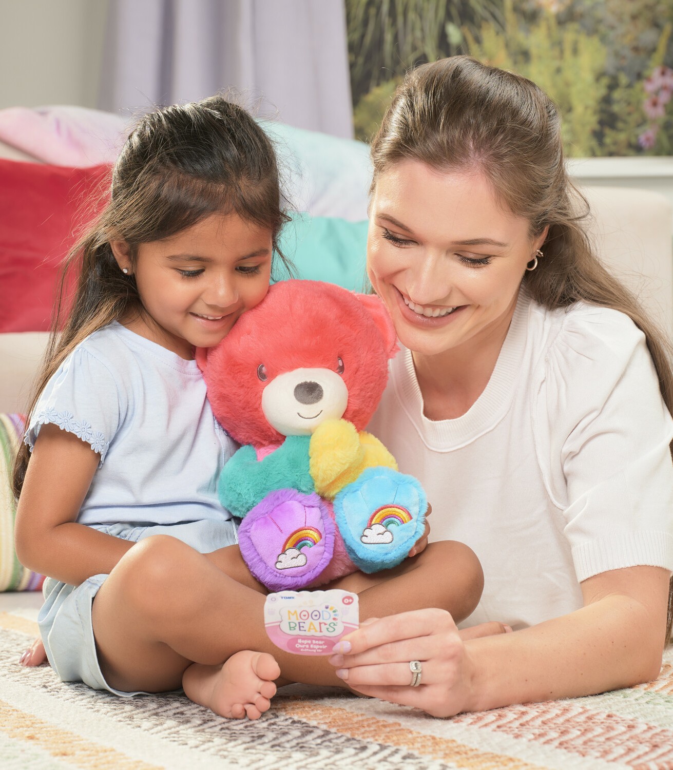 A woman with dark hair and a white t shirt is lying on the floor next to a young girl with dark hair and they are playing with a multi-coloured Mood Bear teddy. They are lying in a bedroom.
