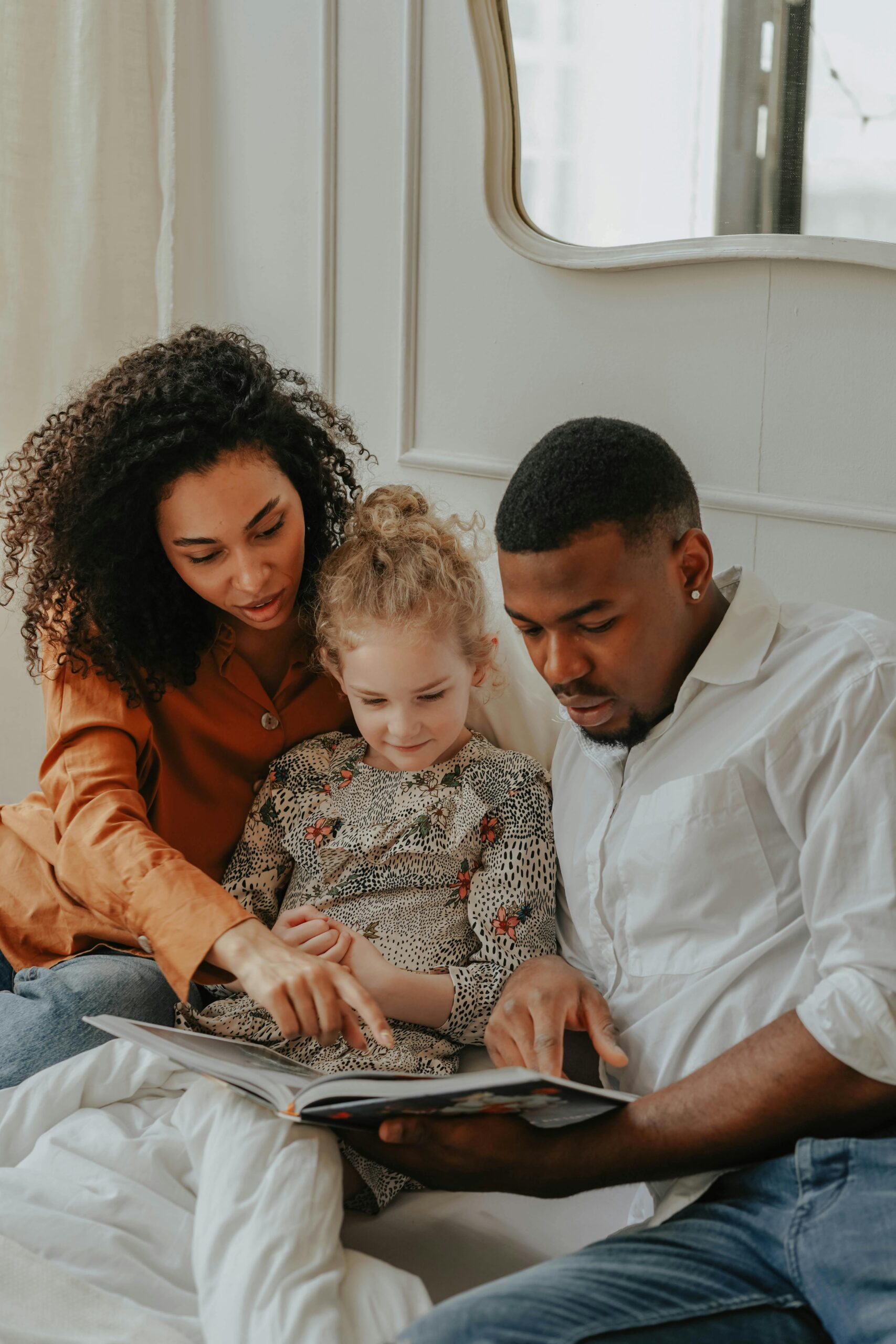 This shows a man woman and child sat on a bed. The woman has curly long dark hair and is wearing a brown top pointing at a book which a young blonde haired girl is holding. On their left is a man sat holding the book in a white shirt and blue jeans.