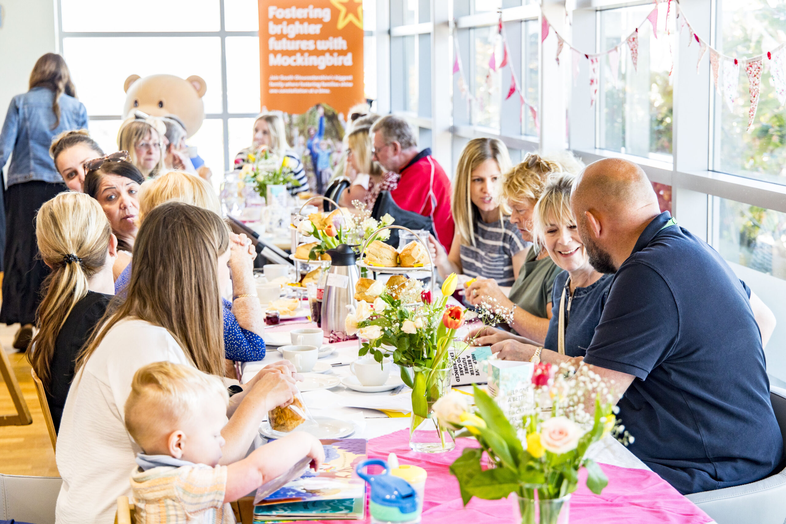 This image captures a warm and welcoming gathering of people seated around a long, flower-adorned table, sharing food and conversation. The setting suggests a community meal or celebration, with a focus on connection and togetherness. In the background, an orange sign reads: "Fostering brighter futures with Mockingbird"