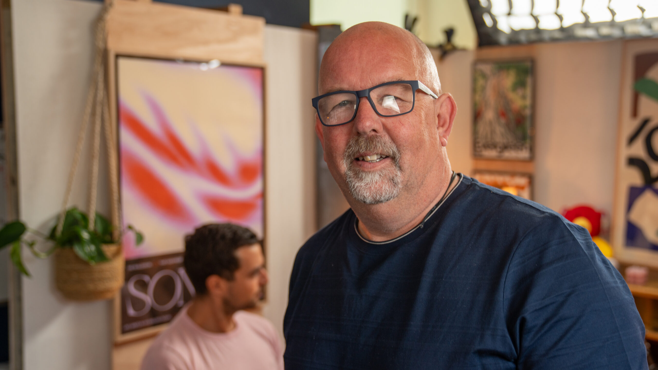 This photo features two individuals in an indoor space with a creative and welcoming atmosphere. The person in the foreground is wearing a dark blue shirt, he has glasses and a facial hair. While the person behind them is dressed in a light pink shirt. The background includes several framed posters and artworks, one of which displays abstract red and white patterns. A hanging plant adds a touch of greenery to the left side of the scene, contributing to the cosy and artistic setting.