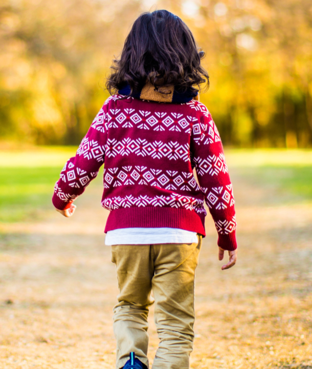 The image shows a young child walking in a park facing away camera. They have dark hair, a dark red jumper and brown trousers.