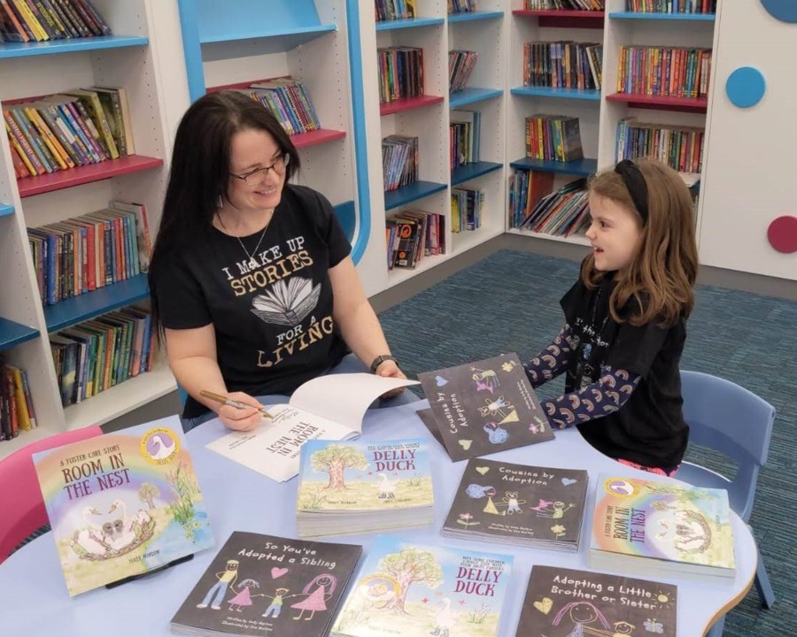 The image shows a woman in a black top with shoulder length dark hair and a young girl with a similar top sat at a table with lots of books. It is taken in a library setting.