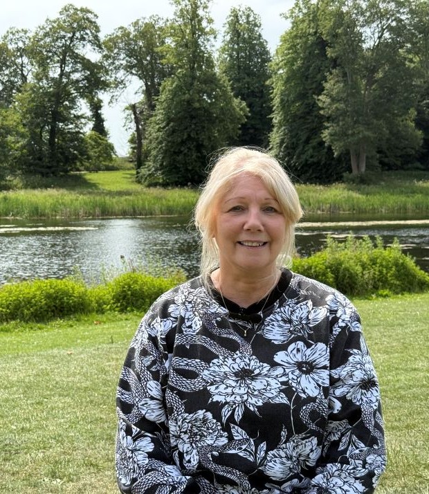 A woman is sat on a bench in a grassy park, behind her you can see a lake and some trees. She is wearing a black and white flowery top and has tied back blonde hair.