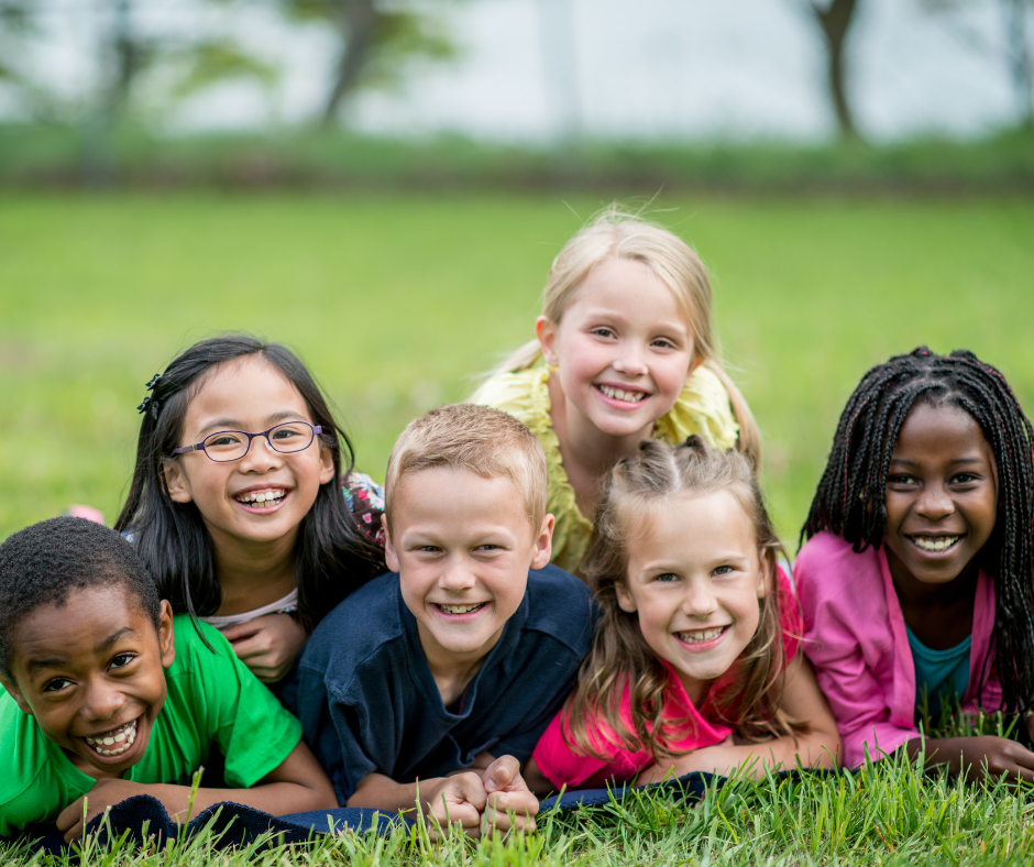 The picture shows six children lying on the grass in an outdoor setting, likely a park or garden. The background features trees and greenery, creating a natural and relaxed atmosphere.
The far left child is wearing a green shirt with short hair.
The second child has long black hair and is wearing a white shirt with a colorful pattern.
The centre child has short blonde hair and is dressed in a dark blue shirt. The fourth child has long light brown hair tied back and is wearing a pink shirt.
Behind the front row, slightly elevated: One child has long blonde hair and is wearing a yellow shirt. The other has long braided black hair and is dressed in a pink jacket over a dark-coloured top. All six children are lying on their stomachs on the grass, positioned closely together, suggesting a relaxed and friendly group setting.