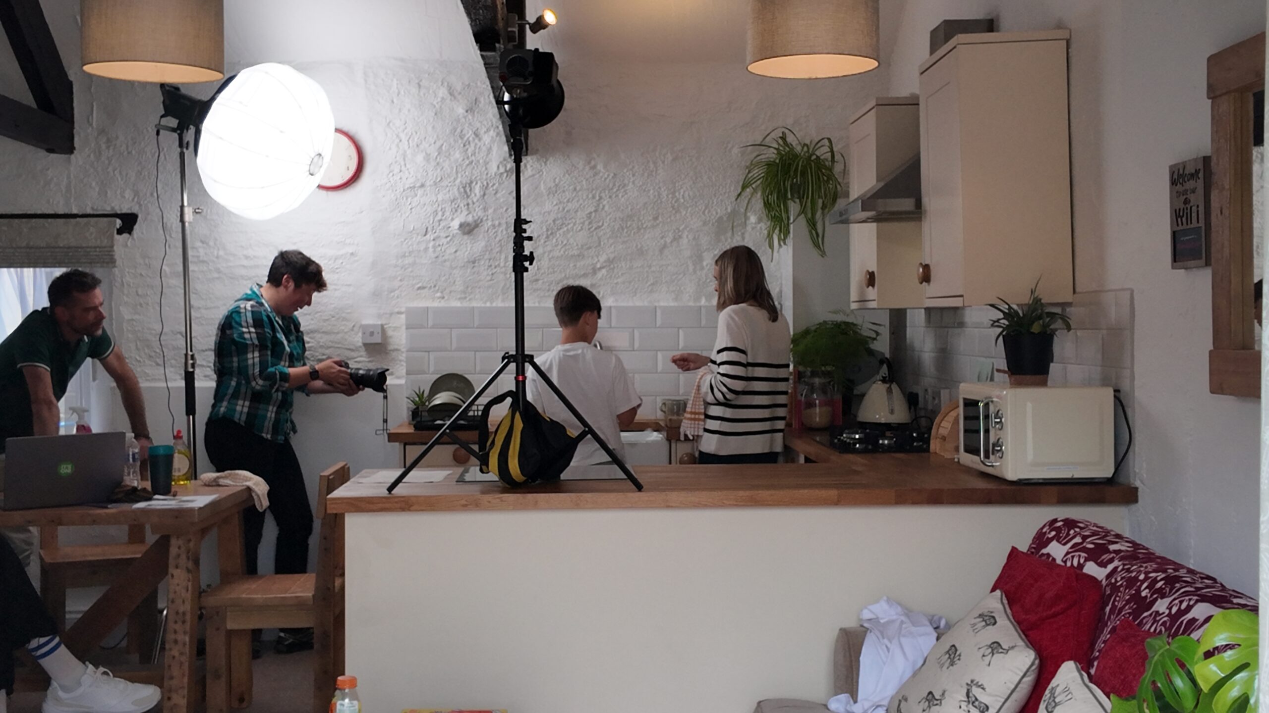 The picture shows a kitchen scene with four people actively engaged in different tasks: Two individuals are standing behind the kitchen counter. One person is washing dishes. Another person is holding a plate. On the left side, there are two more individuals, one of whom are adjusting a camera and lighting equipment, preparing for a photo shoot.
