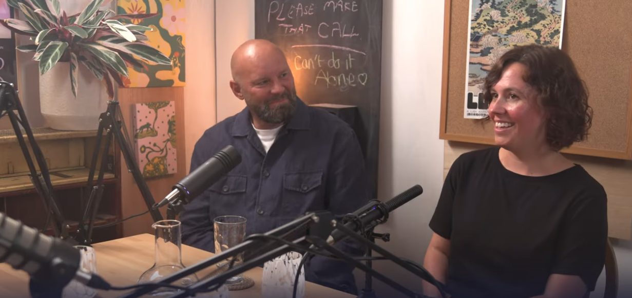 The image captures a warm and engaging podcast setting, featuring two individuals seated at a table with microphones, ready for conversation. The person on the left wears a blue jacket and has a beard, while the person on the right wears a black shirt and has curly hair.