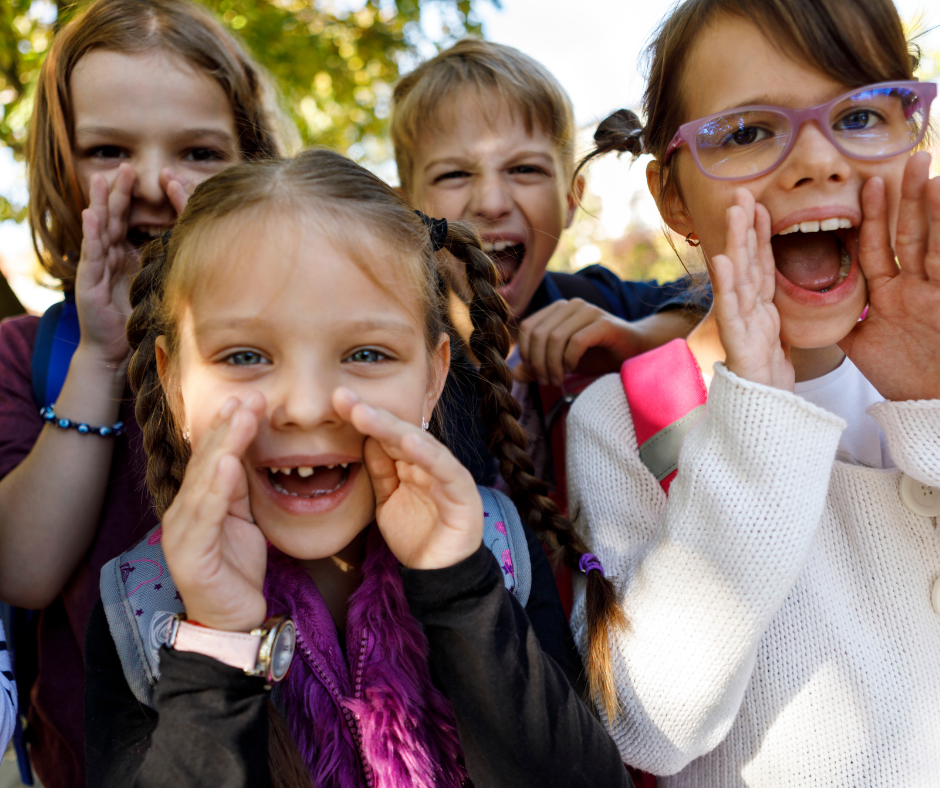 The image features five children standing closely together outdoors, surrounded by trees and bathed in sunlight. Each child is facing the camera with hands cupped around their mouths, as if shouting or calling out. They are dressed in casual clothing, including sweaters and backpacks. The scene conveys a sense of energy and playfulness, capturing a lively moment of interaction among the group.