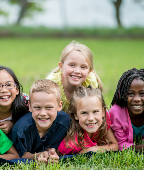 The image shows a group of five children lying on a grassy floor. There is a mixture of boys and girls and they're all smiling at the camera.