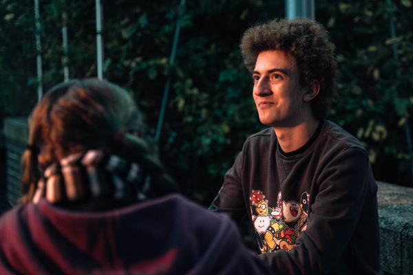 A teenage boy in a dark jumper with curly hair looks away from camera left, whilst a young girl with a black and white scarf faces away from camera and watches him.