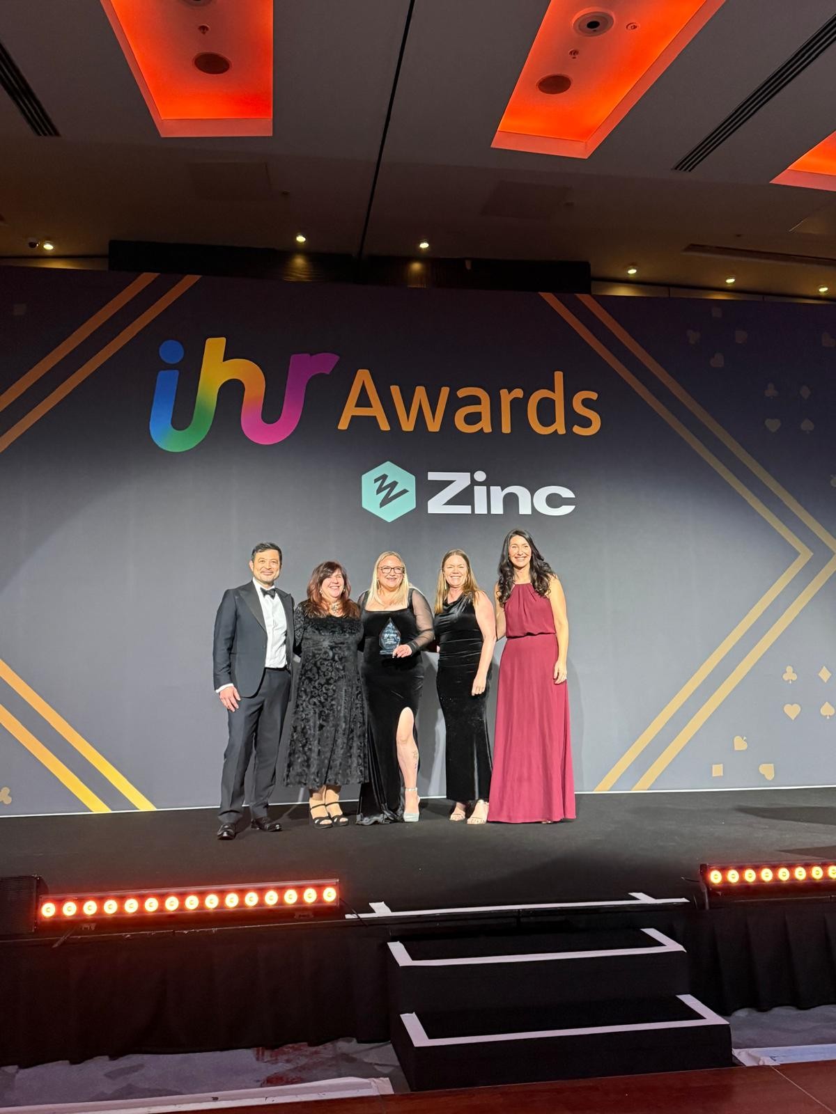 The image shows a group of five people standing on a stage at an awards event. They are dressed in formal evening attire, with three individuals wearing elegant black dresses and one in a deep red gown, while another is in a dark suit. The backdrop features the text “IHR Awards” in large, colorful letters, along with the sponsor logo “Zinc” displayed prominently beneath it. The stage is lit with bright spotlights and has a sleek black floor with a row of red lights along the front edge, creating a celebratory and professional atmosphere. One person in the centre is holding an award, indicating a moment of recognition and achievement.
