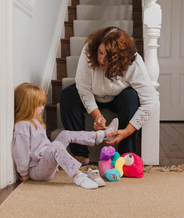 A woman with a cream long sleeve top and dark hair is sat on some stairs in a house helping a young girl in lavender pyjamas tie a shoelace.