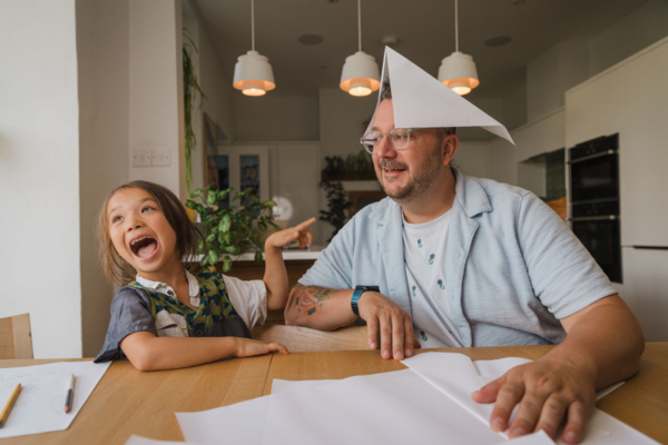 A young girl and man are sat at a kitchen table in a home environment. The young girl in a dark grey top with dark hair is pointing and laughing a the man who has a blue shirt with white undershirt on and has made a paper hat which he is wearing.