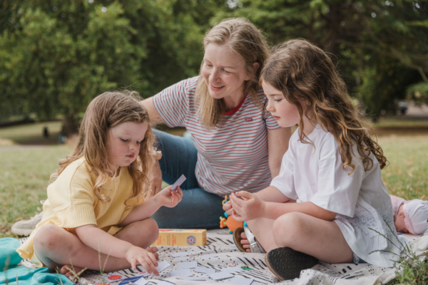A woman and two children are sat in a park on a rug. The youngest girl on the left has brown hair and holding some paper in her hand, the older child on the right is playing with something in her hands and the woman in a striped top with long dark hair watches on.