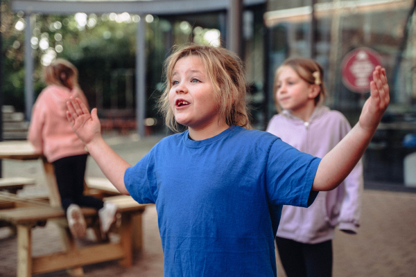 In the foreground of the image is a young girl with a blue shirt an light brown hair with her arms out. Behind her is a young girl in a lilac jumper looking off camera and another young girl with a peach hoodie and dark trousers kneeling on a bench looking away from camera.