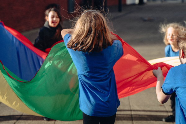 The image shows a group of children playing with a multi coloured parachute and bouncing a toy on it.