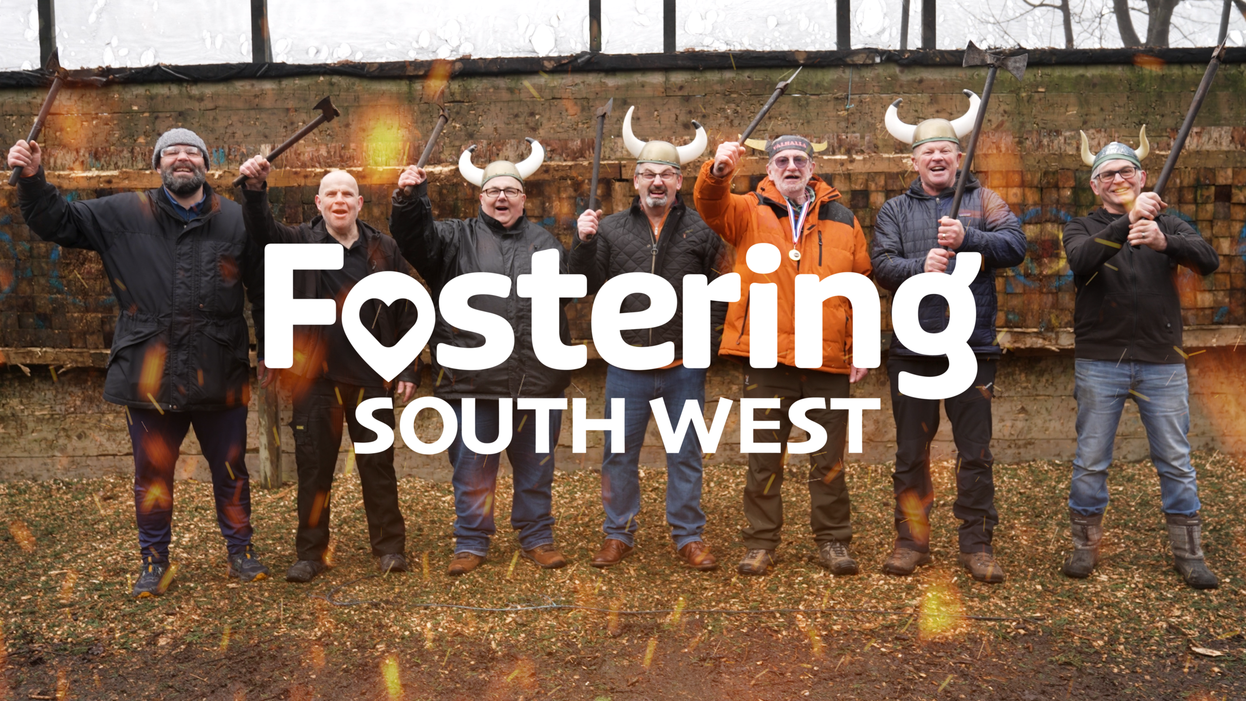 The image shows a group of people standing in a line outdoors at what appears to be an axe‑throwing activity. Each person is holding an axe raised in the air, and several are wearing playful Viking‑style horned helmets. They are standing in front of a rustic wooden target wall, with scattered woodchips on the ground. The Fostering South West logo is prominently displayed across the centre of the image. The overall atmosphere is energetic and fun, suggesting a team‑building or social event.